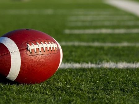 A close-up of a red and white football resting on green turf near the white yard line, with a blurred field in the background, on a sunny day.