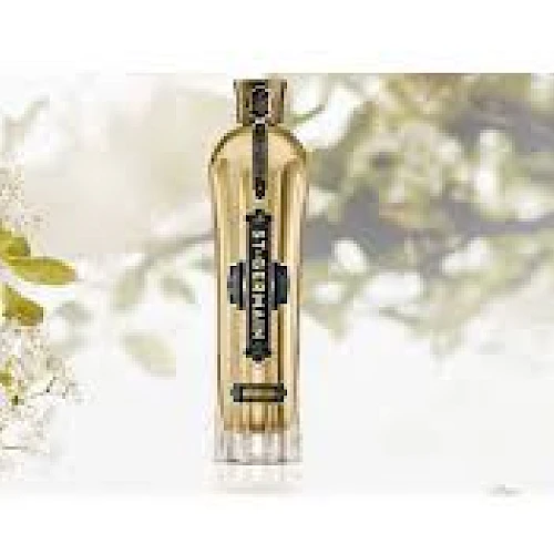 A bottle of perfume or fragrance stands upright against a light, floral background, framed by soft white flowers and greenery, sunlit.