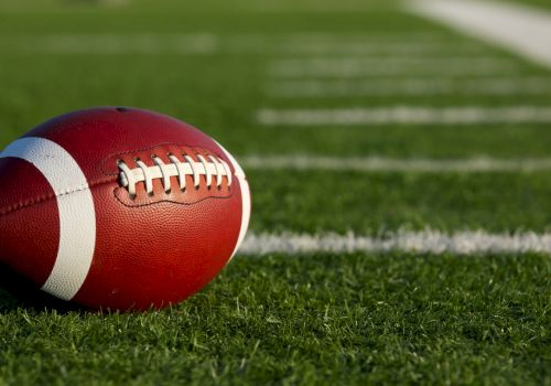 A red and white American football rests on green turf near the sideline, ready for play.