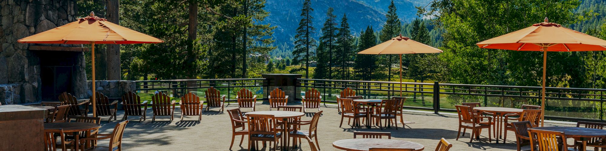 An outdoor patio area features several wooden tables and chairs, each with orange umbrellas. It is surrounded by trees and mountains, under a clear sky.