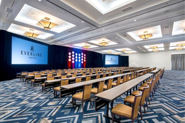 A spacious conference room with rows of tables and chairs facing multiple large screens displaying 