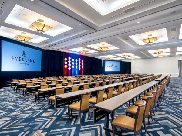 A spacious conference room with rows of tables and chairs facing multiple large screens displaying "Everline." The room has modern lighting.