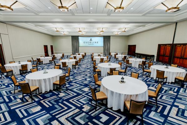 A spacious conference room with round tables covered in white cloth, each surrounded by chairs. There is a large screen displaying 