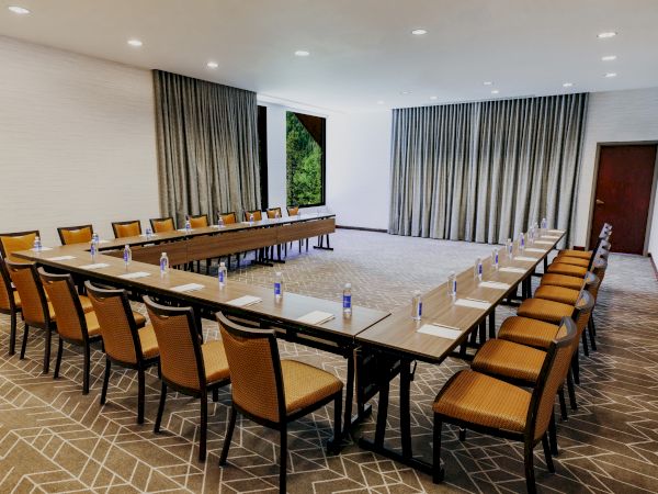 A conference room set up with U-shaped tables, chairs, notepads, and water bottles, with curtains and a door in the background ends the sentence.