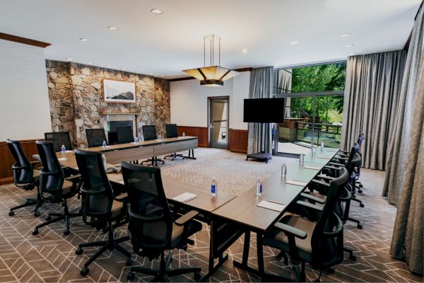 A modern conference room with a long table, multiple chairs, a TV, and large windows with a view outside; water bottles and notebooks on the table.