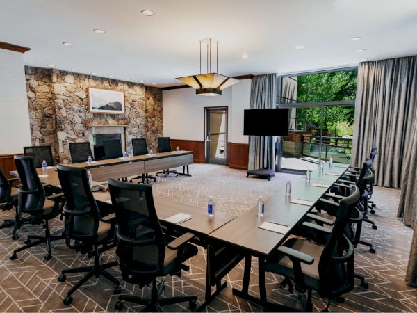 A modern conference room with a long table, multiple chairs, a TV, and large windows with a view outside; water bottles and notebooks on the table.
