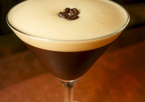 A frothy coffee cocktail in a coupe glass, dark espresso base topped with a creamy foam and a coffee bean garnish.