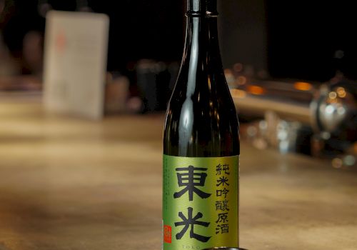 A bottle of sake with a green label sits on a bar counter, two small decorated cups beside it, warm ambient lighting.