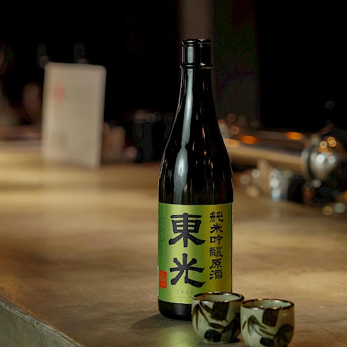 A bottle of sake with a green label sits on a bar counter, two small decorated cups beside it, warm ambient lighting.