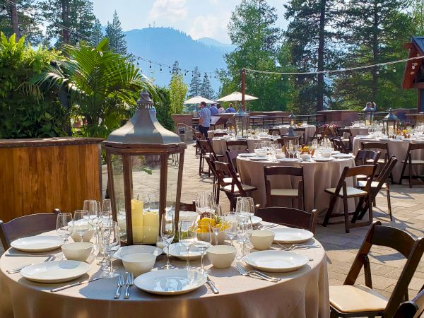 An elegantly set outdoor dining area with round tables, wooden chairs, a lantern centerpiece, and a mountain scenery in the background.