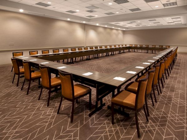 The image shows a U-shaped conference room setup with rows of chairs and tables aligned, with notepads placed on the tables, and a carpeted floor.