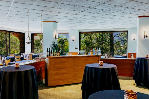 The image shows a modern dining area or event space with round tables covered in dark cloths, a wooden bar, and large windows with a view of greenery outside.