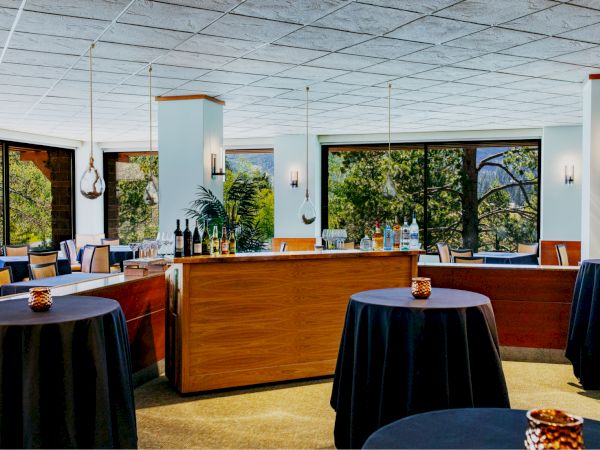 The image shows a modern dining area or event space with round tables covered in dark cloths, a wooden bar, and large windows with a view of greenery outside.