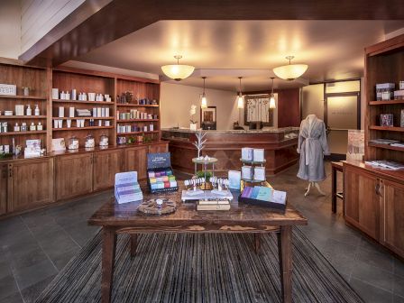 A boutique apothecary or spa shop with wooden shelves lined with jars and bottles, a central display table, and a person browsing in a robe.