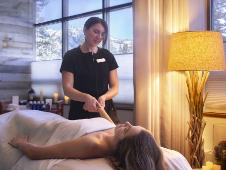 A spa therapist stands beside a client lying on a massage table, preparing oils in a calm, softly lit treatment room.