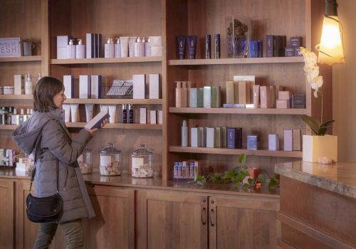 A woman browsing skincare products on wooden shelves in a store, selecting items to inspect.