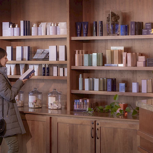 A woman browsing skincare products on wooden shelves in a store, selecting items to inspect.