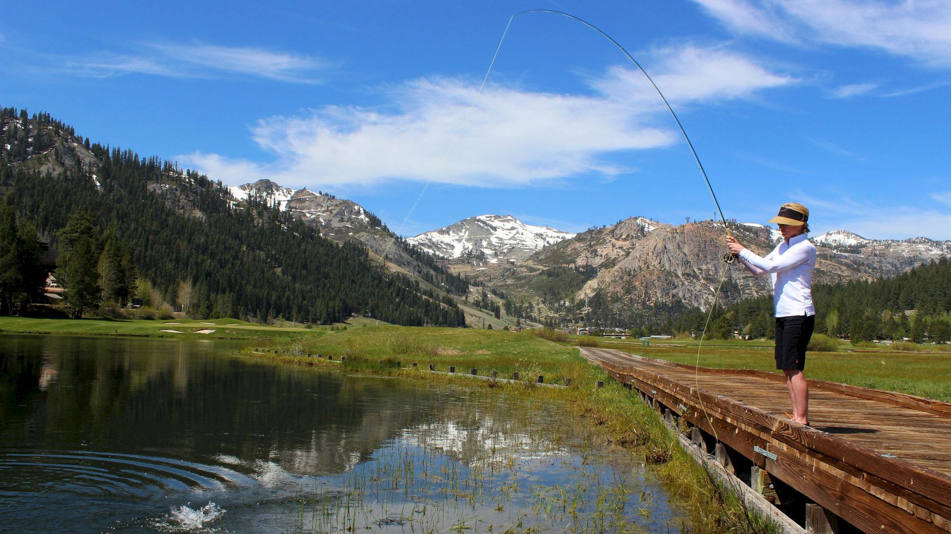 A person is fishing from a wooden dock surrounded by mountains and forest, with clear blue skies and a scenic lake reflecting the landscape.