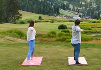Two people stretch on yoga mats on a grassy hill, facing a scenic valley in the distance.