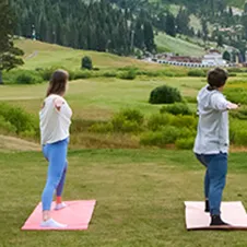 Two people stretch on yoga mats on a grassy hill, facing a scenic valley in the distance.