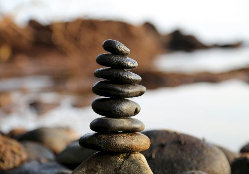 A balanced stack of smooth, dark stones stacked on a beach, with a blurred rocky shoreline in the background.