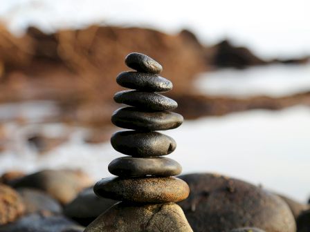 A balanced stack of smooth, dark stones stacked on a rocky shore by the water, with a calm, blurred background.