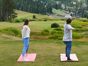 Two people stand on yoga mats in a grassy meadow, stretching and looking out over green hills in nature.