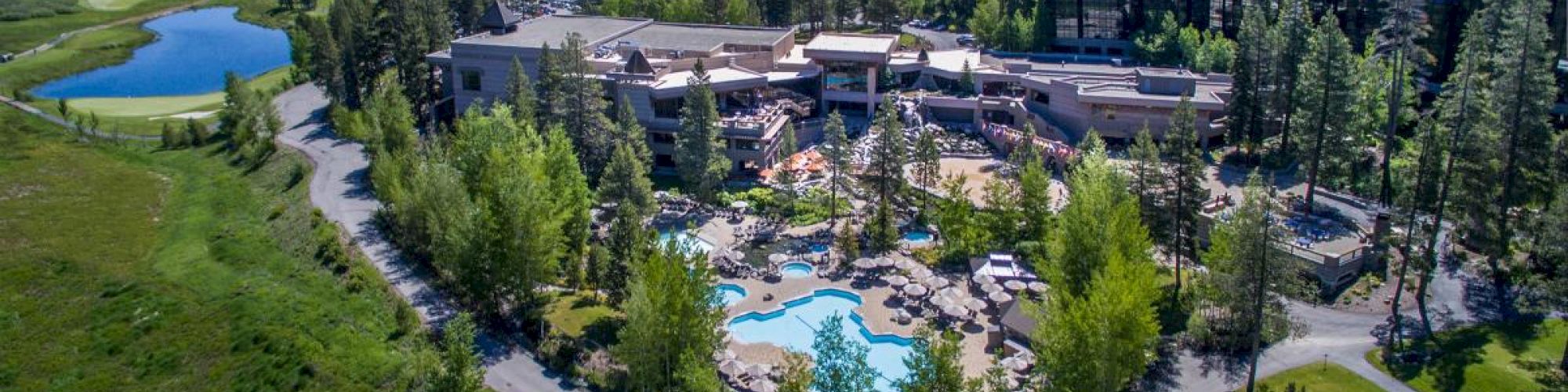 The image shows an aerial view of a resort nestled in a forested area with mountains in the background, featuring a golf course, swimming pool, and various buildings.