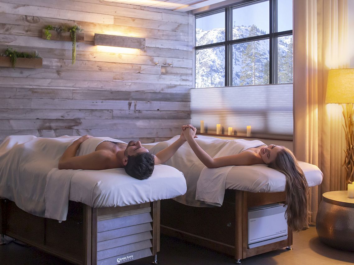 Two people lie on massage tables in a spa, holding hands and smiling, while a serene, softly lit room surrounds them.