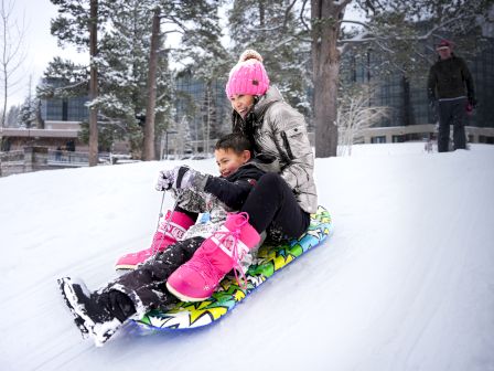 Two people sledding down a snowy hill, wearing winter clothes and pink boots, with trees and a building in the background.