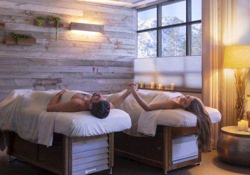Two people lie on separate massage beds in a spa, holding hands and facing each other, relaxing in a cozy, softly lit room.