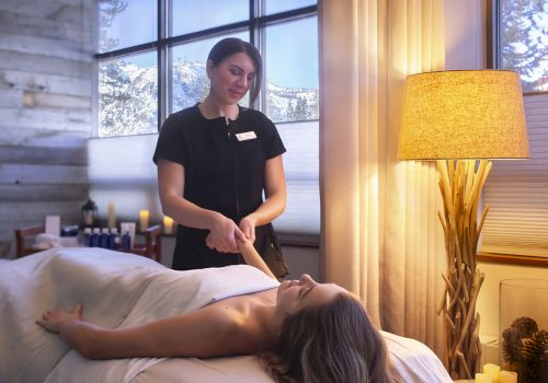 A spa therapist prepares to treat a client lying face up on a massage table in a cozy, softly lit room with a lamp and windows.