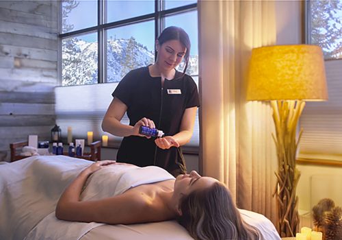 A massage therapist works on a client lying face up on a massage table, with oils and a lamp in a softly lit spa setting.
