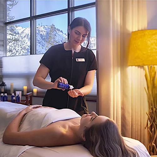 A massage therapist works on a client lying face up on a massage table, with oils and a lamp in a softly lit spa setting.