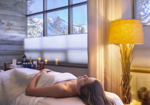 A woman relaxes in a spa-like room, lying on a massage bed with soft lighting, candles, and a warm lamp by the window.