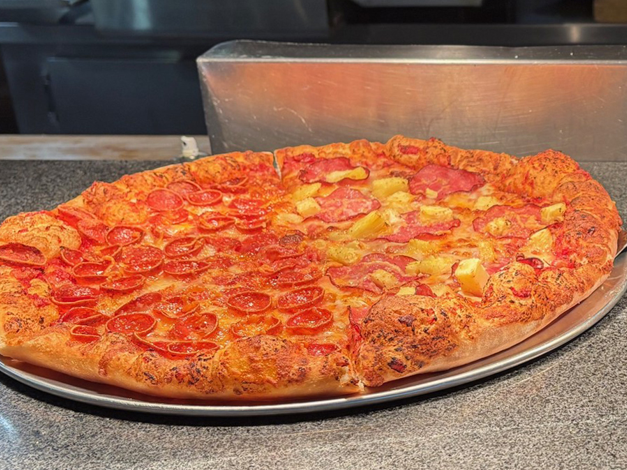 A large pepperoni and pineapple pizza on a metal tray, with a golden, bubbling crust at a pizza counter.