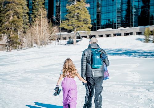 Two people, a child and an adult, walk hand-in-hand through a snowy path near a modern glass building, with pine trees and blue skies overhead.