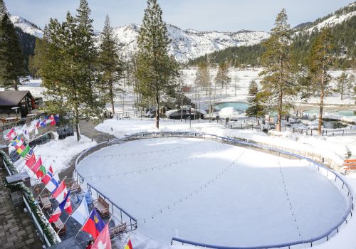 A snowy lakeside scene with a circular ice rink, flags along the edge, pine trees, and mountains in the background.