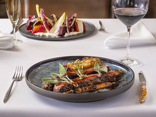 A fine dining plate with seared meat, asparagus, and greens, plus a plated beet salad in the background, wine glasses, and cutlery, on a white tablecloth.