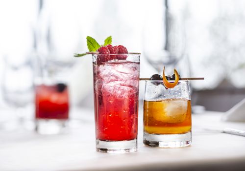 Two colorful cocktails on a bright table, garnished with mint and fruit, one red and one amber, with a blurred drink in the background.