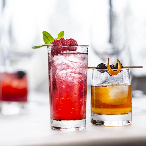 Two colorful cocktails on a bright table, garnished with mint and fruit, one red and one amber, with a blurred drink in the background.
