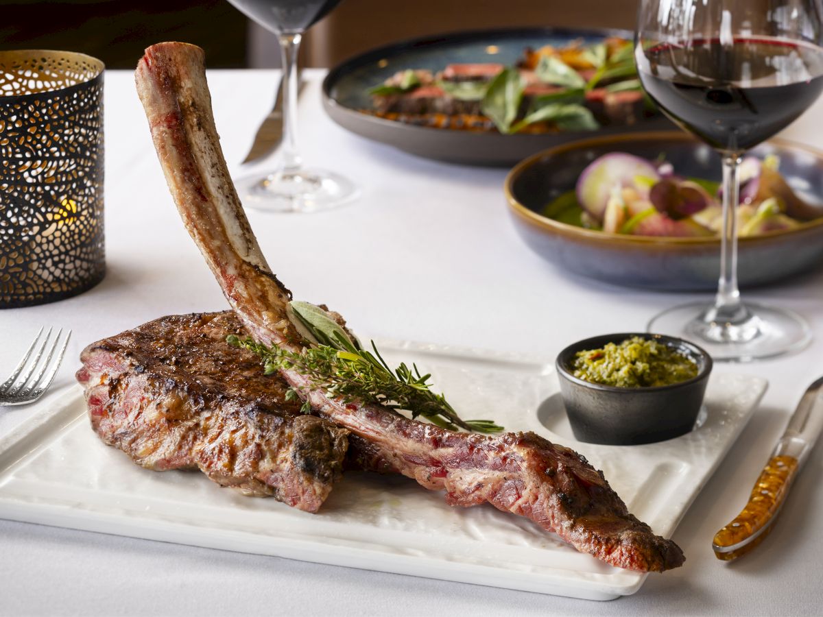 A plate with a large tomahawk steak garnished with herbs, a small bowl of sauce, a candle, wine glasses, and additional dishes in the background.