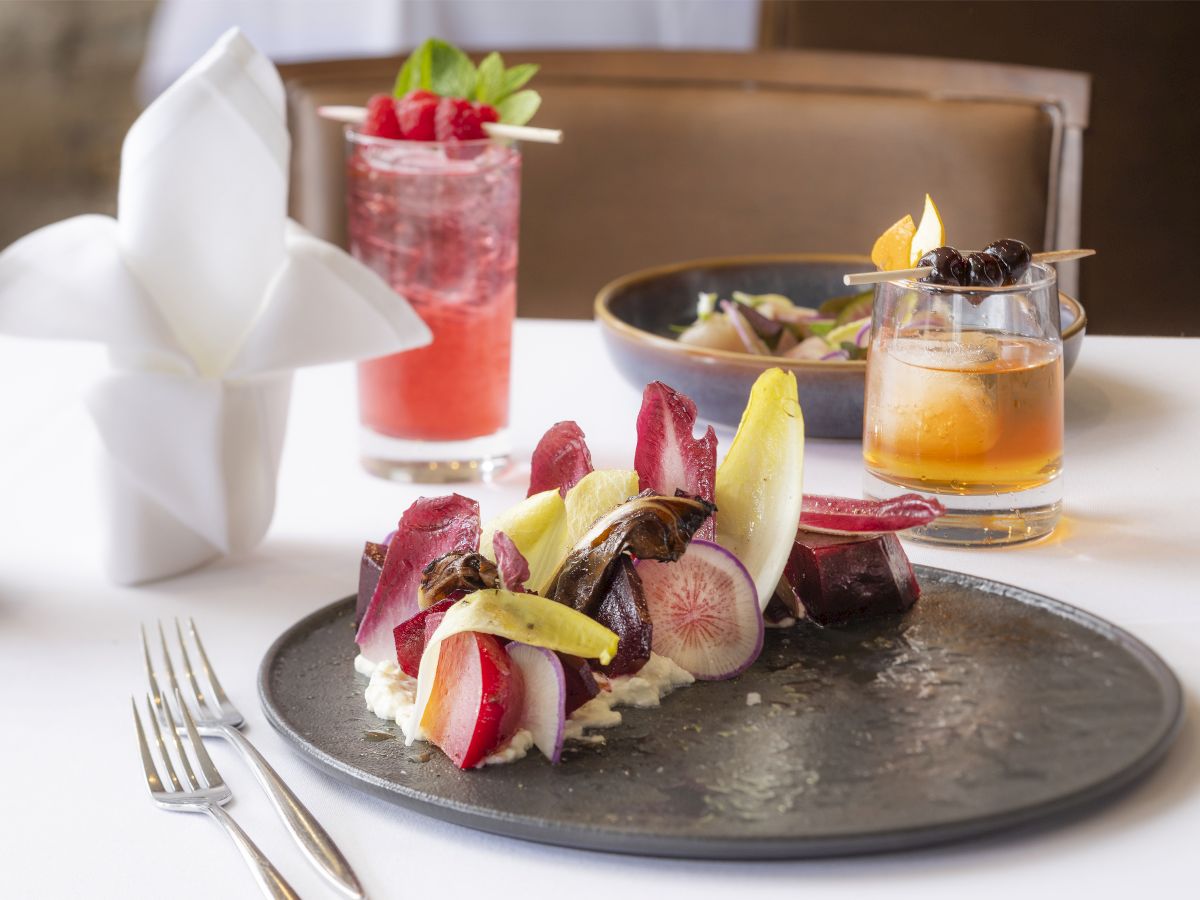 A beautifully plated dish with colorful vegetables, two cocktails, and a napkin folded elegantly, set on a white tablecloth.