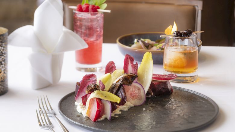 A beautifully plated dish with colorful vegetables, two cocktails, and a napkin folded elegantly, set on a white tablecloth.