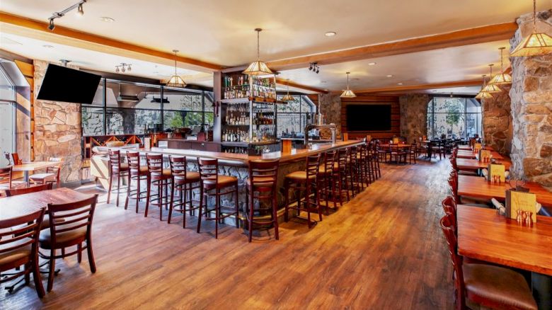 The image shows an empty restaurant with wooden tables and chairs, a central bar area, stone walls, and large windows providing natural light.