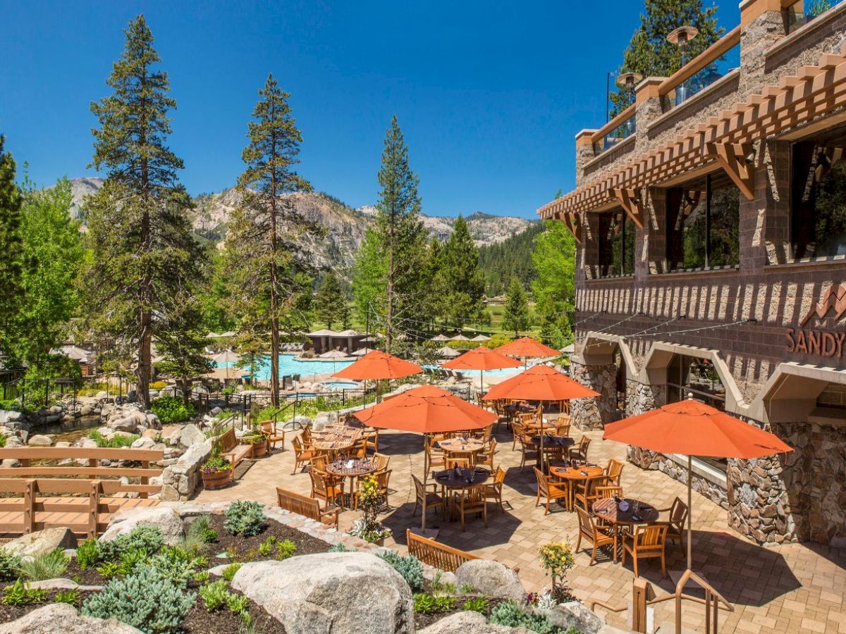 A scenic restaurant with outdoor seating and orange umbrellas, surrounded by trees and mountains, with a pool in the background.