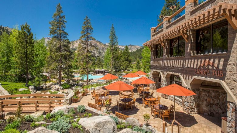 A scenic restaurant with outdoor seating and orange umbrellas, surrounded by trees and mountains, with a pool in the background.