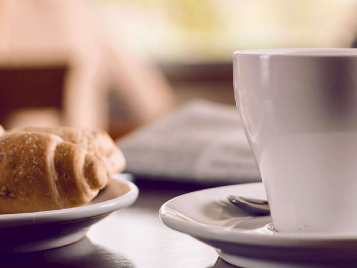 A white coffee cup on a saucer sits next to a plate of pastries on a table, with a rolled-up newspaper in the background.