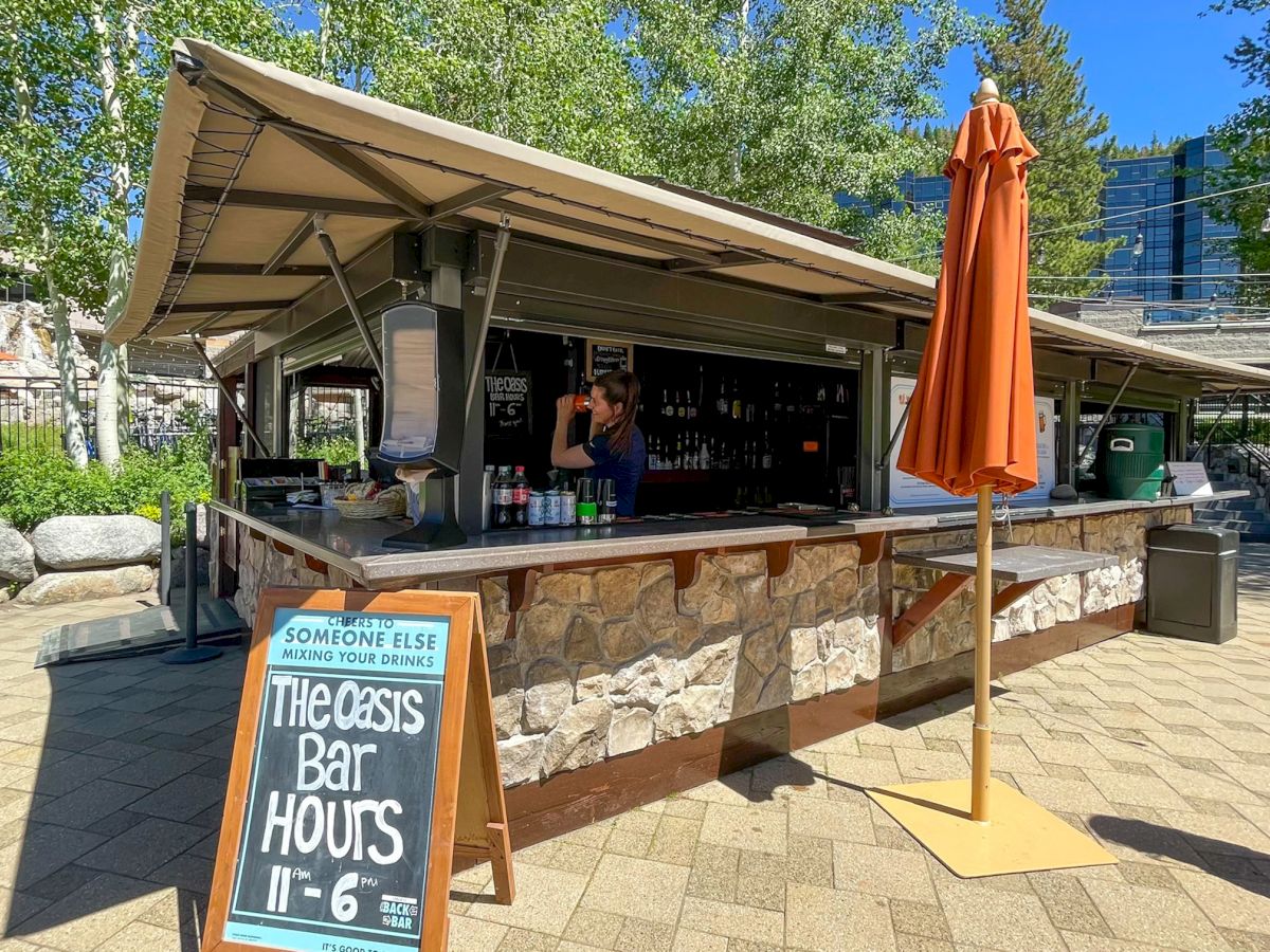 An outdoor bar with a stone facade and awning, featuring a chalkboard sign with hours, various drinks, and an orange umbrella. The end.