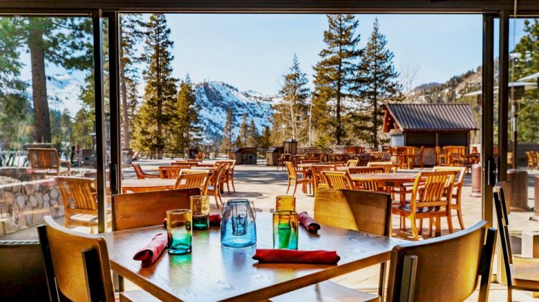 A restaurant patio with colorful glasses, napkins, wooden tables and chairs, overlooking a scenic view of trees and snow-capped mountains.